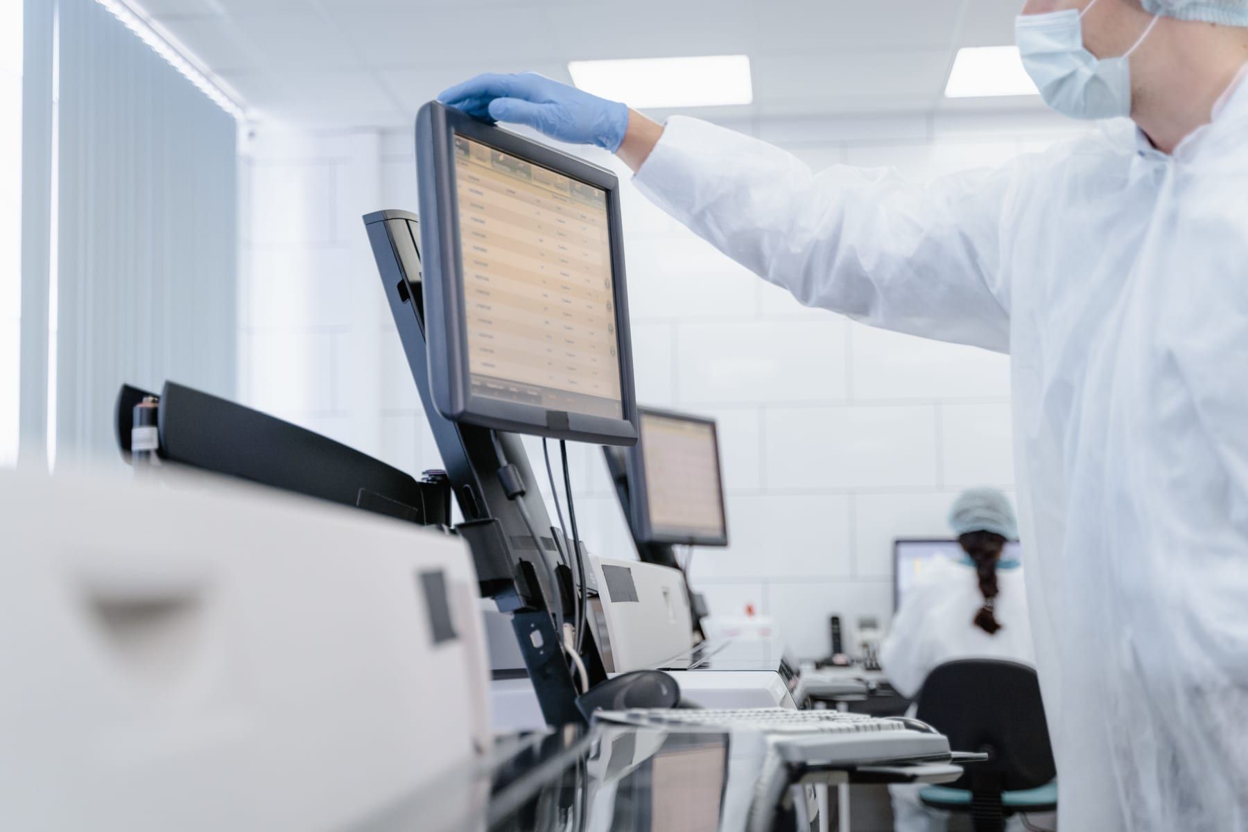 An analyst in a gown and gloves checks a results table on a screen mounted beside the instrument.