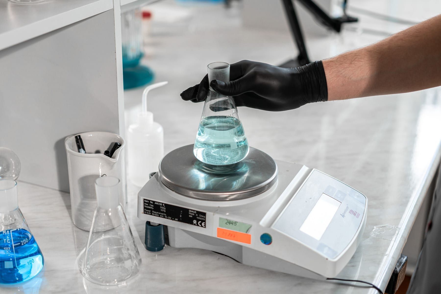 An analyst in black gloves sets a flask of solution on an analytical balance.