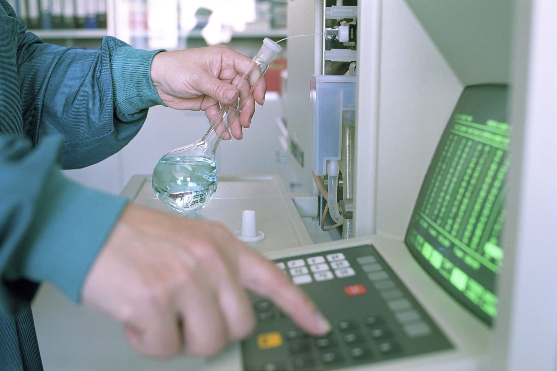 An analyst holds a flask and keys a command into an instrument panel with a green readout.